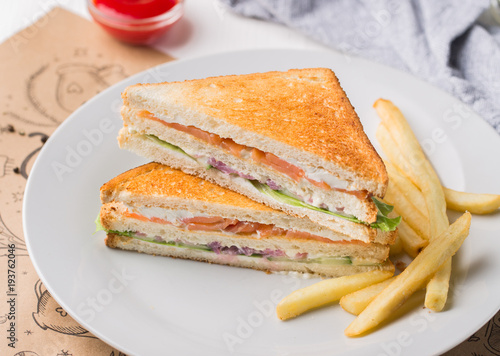 Freshly made club sandwiches with French fries served on white plate. Pressed double toasts with leaf vegetable salad, smoked salmon and cheese served on paper on a wooden table. Street fast food.