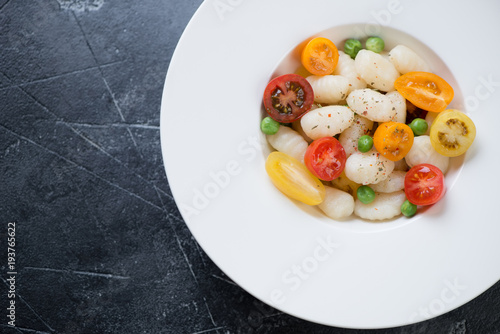 White plate with italian gnocchi, fresh tomatoes and green peas, view from above on a grey concrete background, horizontal shot with space