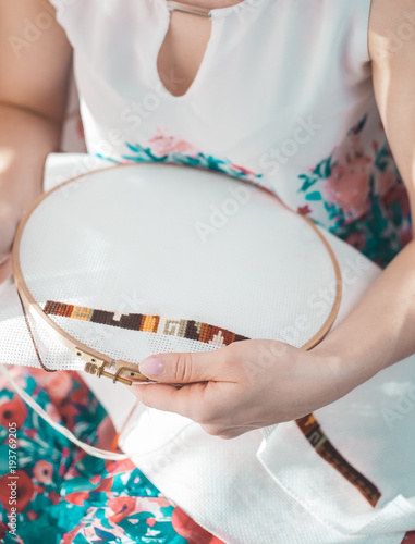 Young beautiful woman is engaged in embroidering a dagger on the embroidery frame in a colored dress at the window in the sunlight. A great picture for a hobby