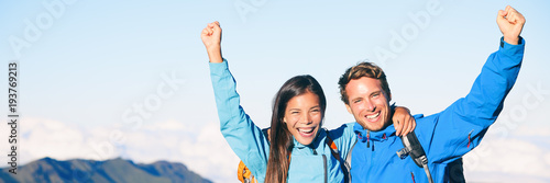 Happy mountain climbers hiking mountains reaching mountain summit cheering with arms up in success. Successful tourists couple trekking in altitude banner panorama.