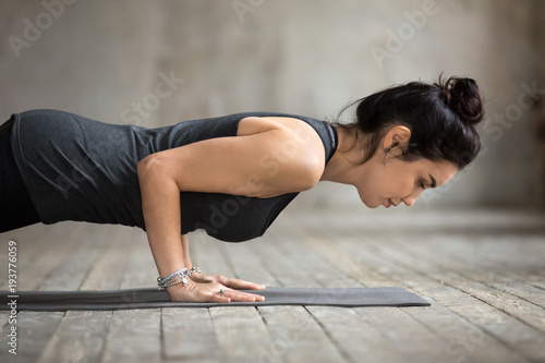 Young woman practicing yoga, doing four limbed staff, Push ups or press ups, exercise, chaturanga dandasana pose, working out, wearing sportswear, black tank top, close up, gray wall in yoga studio