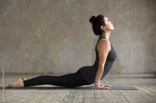 Young woman practicing yoga, doing upward facing dog exercise, Urdhva mukha shvanasana pose, working out, wearing sportswear, black pants and top, indoor full length, gray wall in yoga studio