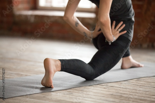 Young woman practicing yoga, doing Horse rider exercise, anjaneyasana pose, working out, wearing sportswear, black pants and top, indoor close up, gray wall in yoga studio