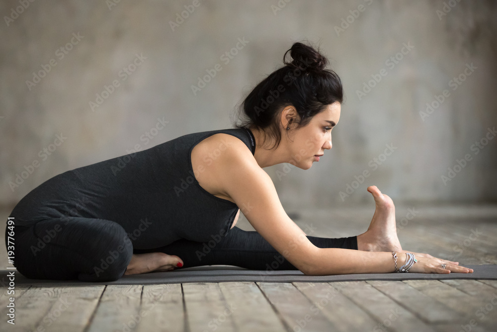 Young woman practicing yoga, doing Head to Knee Forward Bend exercise, Janu Sirsasana pose, working out, wearing sportswear, black pants and top, indoor full length, gray wall in yoga studio Young woman practicing yoga, doing Head to Knee Forward Bend exercise, Janu Sirsasana pose, working out, wearing sportswear, black pants and top, indoor full length, gray wall in yoga studio