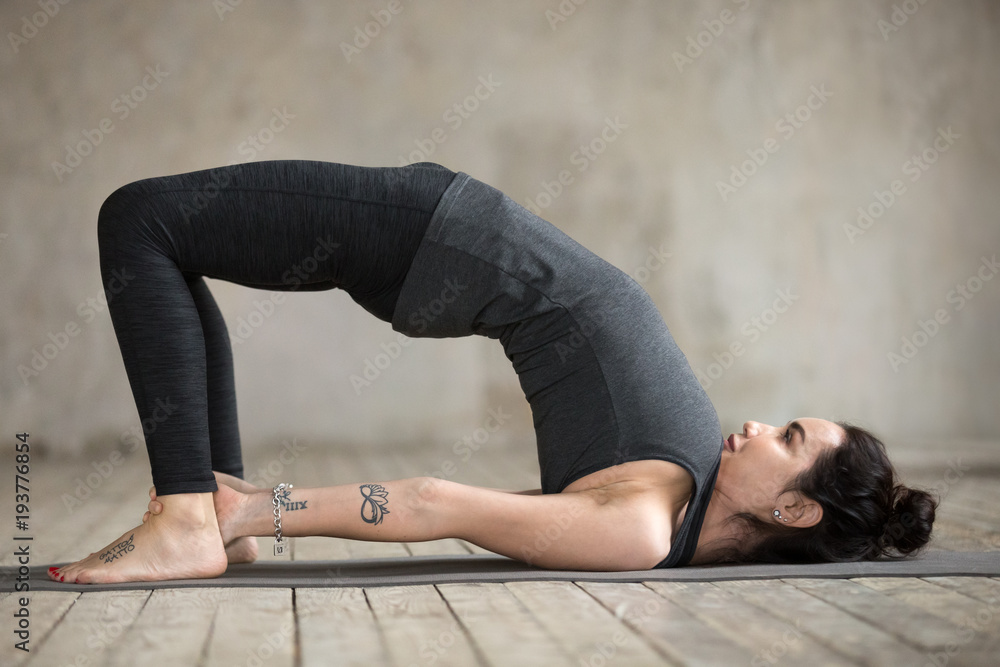 Obraz premium Young sporty woman practicing yoga, doing dvi pada pithasana exercise, Glute Bridge pose, working out, wearing sportswear, black pants and top, indoor full length, gray wall in yoga studio