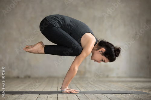 Young woman practicing yoga, doing Crane exercise, Bakasana pose, working out, wearing sportswear, black pants and top, indoor full length, gray wall in yoga studio