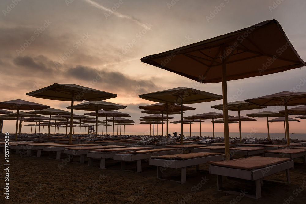 Silhuettes of beach loungers and umbrellas on empty beach in evening on a sunset background
