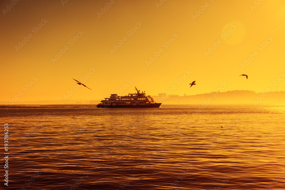 Silhouette of a ferry boat at sunset and seagulls fly around. Beautiful atmospheric bright landscape