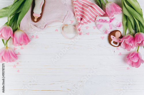 Fototapeta Naklejka Na Ścianę i Meble -  A composition for newborns on a wooden white background with clothes, pink tulips, hearts and a cookies, copy space and flat lay. It's a girl.
