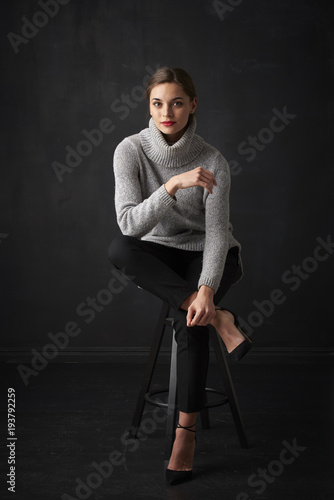 Relaxed young woman at dark background. Full length shot of attractive young woman wearing turtleneck sweater while sitting at dark background and relaxing. 
