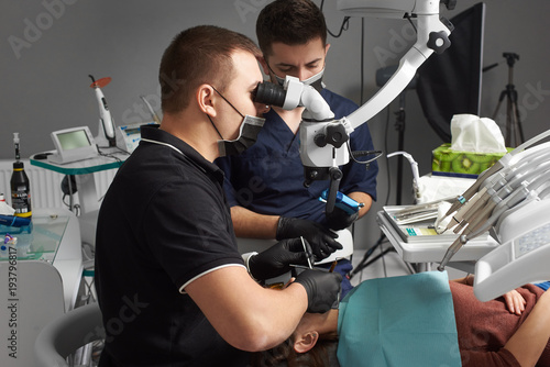 Male dentist with an assistant in a black mask and gloves treats a patient using a microscope. Modern dentistry with the use of new technologies