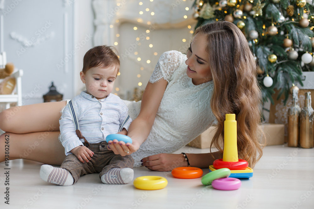 Naklejka premium Mother playing with her son on the eve of the New Year. Mother teaches young son to collect the multicolored pyramid. Happy family on Christmas tree background.