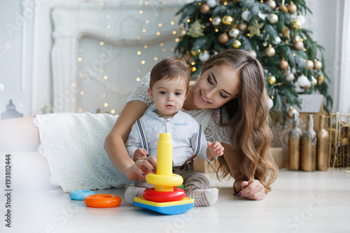 Mother playing with her son on the eve of the New Year. Mother teaches young son to collect the multicolored pyramid. Happy family on Christmas tree background.