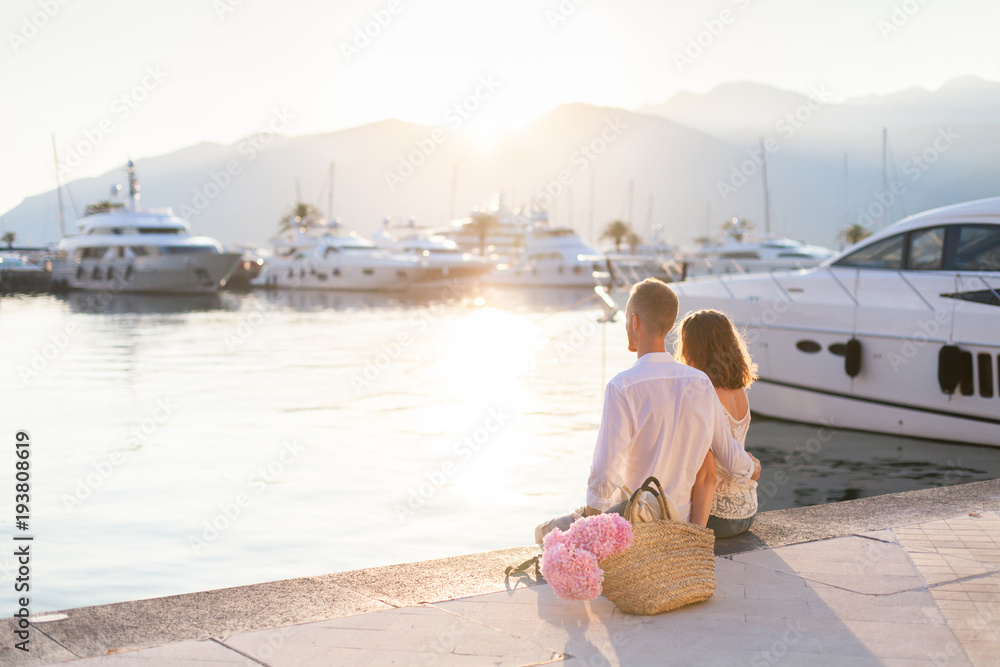 Couple of travelers are sitting in sea yacht port during romantic honeymoon vacation at sunset. Man and woman are tourists. Lovers have pink hydrangea flowers in straw bag and hat outdoor.