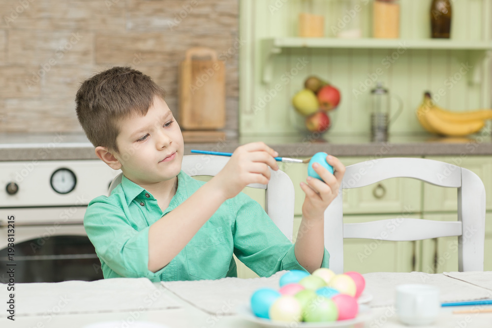 Fototapeta premium Little blond kid boy coloring eggs for Easter holiday in domestic kitchen