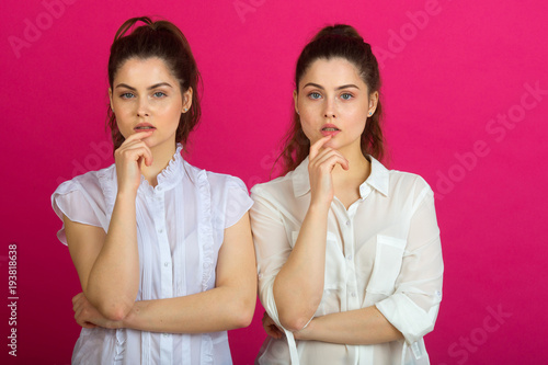 two beautiful young twin sisters in a good mood in white blouses on a pink background with a pensive expression