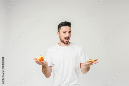Handsome guy is stading at the white wall and holding a small apple in one hand and tasty hamburger in another. He is looking straight with a sight full of excitement and want to ask you a question. photo