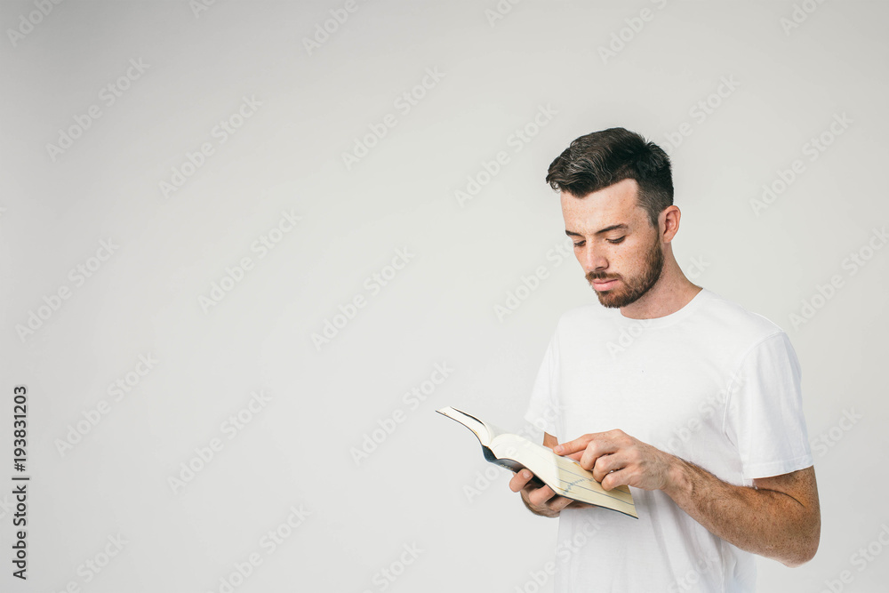 Horizontal photot of a young man standing near the white wal and reading a book. It is so interesting he cannot turn his eyes from it. Horizontal photot of a young man standing near the white wal and reading a book. It is so interesting he cannot turn his eyes from it.
