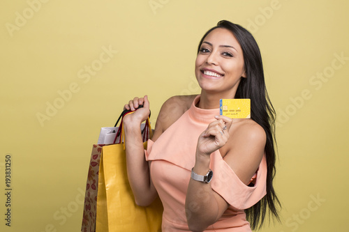 Portrait of a satisfied happy girl holding shopping bags and showing credit card while looking at camera isolated over yellow background
