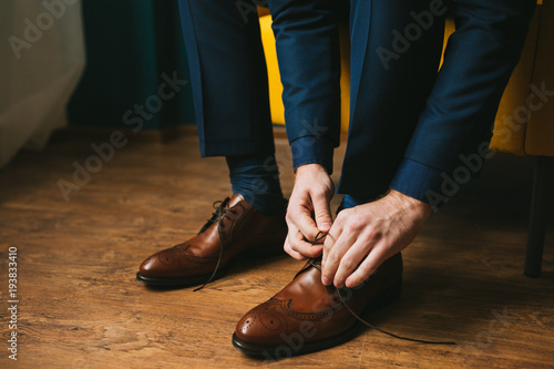 Fotografía A man or a groom in a blue suit ties up shoelaces on brown leather shoes brogues