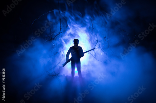 Man with riffle at spooky forest at night with light, or War Concept. Military silhouettes fighting scene on war fog sky background, World War Soldier Silhouette Below Cloudy Skyline At night.