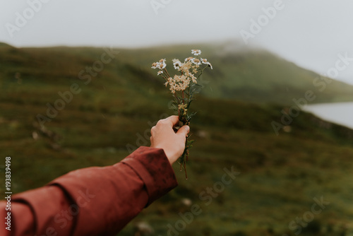 Woman wearing a red rain jacket holding a flower bouquet in front of a green field bokeh unsharp background in Scotland, UK