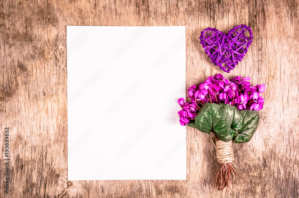 Obraz premium The first spring flowers and wicker heart on a wooden board. Flowers and blank sheet of paper. Romantic concept.