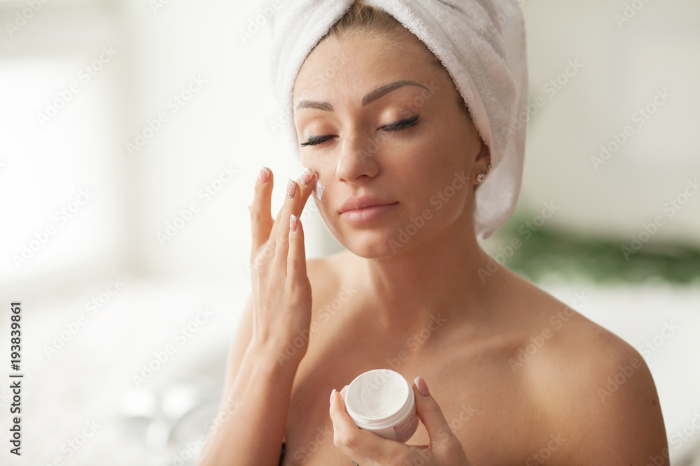 Young woman with flawless skin, applying moisturizing cream on her face. Photo of woman after bath in white bathrobe and towel on white background. Skin care concept Young woman with flawless skin, applying moisturizing cream on her face. Photo of woman after bath in white bathrobe and towel on white background. Skin care concept