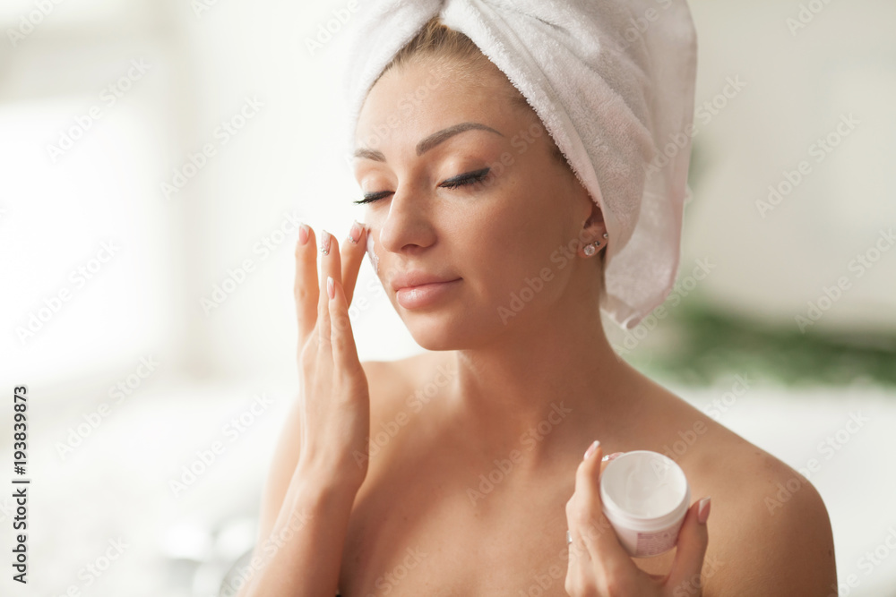 Young woman with flawless skin, applying moisturizing cream on her face. Photo of woman after bath in white bathrobe and towel. Skin care concept Young woman with flawless skin, applying moisturizing cream on her face. Photo of woman after bath in white bathrobe and towel. Skin care concept