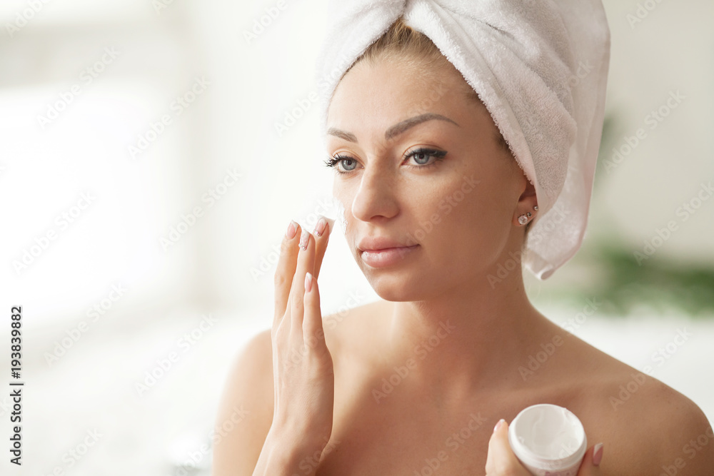 Young woman with flawless skin, applying moisturizing cream on her face. Photo of woman after bath in white bathrobe and towel. Skin care concept Young woman with flawless skin, applying moisturizing cream on her face. Photo of woman after bath in white bathrobe and towel. Skin care concept
