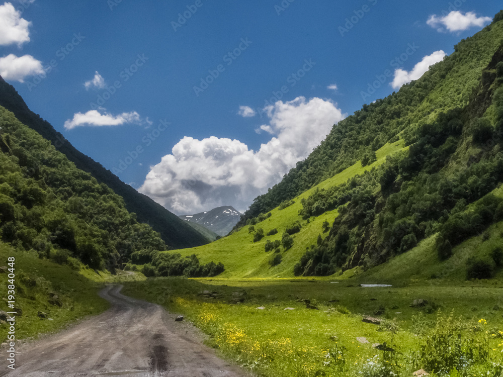 Hot summer day at the valley at Caucasus mountains (Hevsureti, Georgia). Road surrounded with high hills covered with green grass and forests. There are a couple of clouds in the deep blue sky. Hot summer day at the valley at Caucasus mountains (Hevsureti, Georgia). Road surrounded with high hills covered with green grass and forests. There are a couple of clouds in the deep blue sky.
