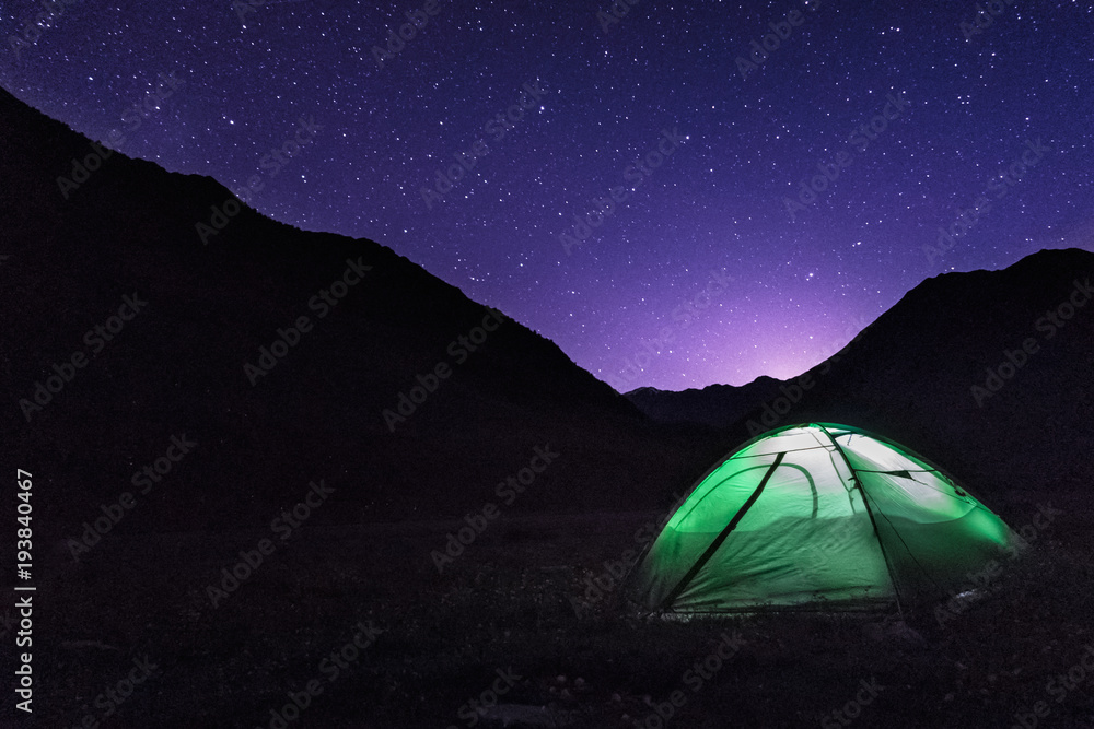 Astrophotography of night camp and Milky way. Dark night and bright galaxy above Caucasus mountains in Georgia. Green tent on the foreground is highlighted from the inside. Backpacking lifestyle.