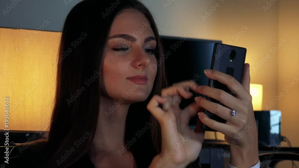 A young beautiful woman works on a smartphone in a dimly lit room - closeup A young beautiful woman works on a smartphone in a dimly lit room - closeup