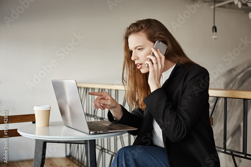 Girl picking item in online-store. Indoor shot of good-looking female student in cafe, sitting over laptop, pointing at screen while talking on smartphone, wanting to buy something in internet