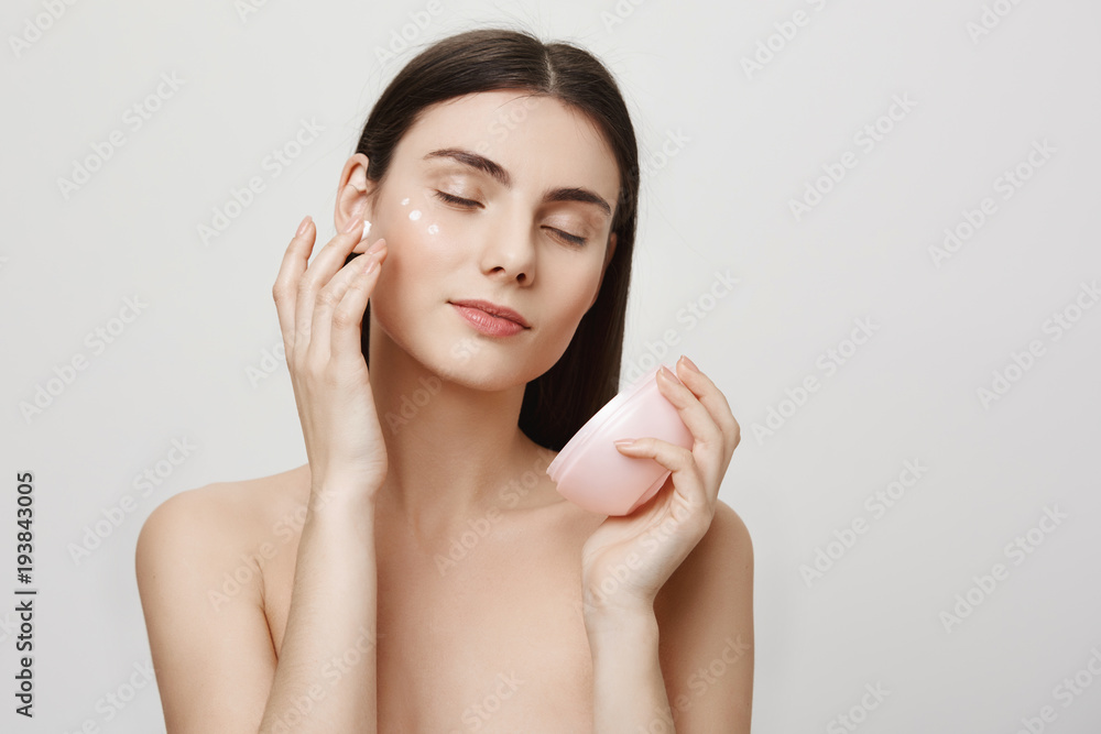 Beauty and skincare concept. Studio portrait of gentle good-looking european woman rubbing in facial cream, enjoying doing cosmetological procedures while standing over gray background Beauty and skincare concept. Studio portrait of gentle good-looking european woman rubbing in facial cream, enjoying doing cosmetological procedures while standing over gray background