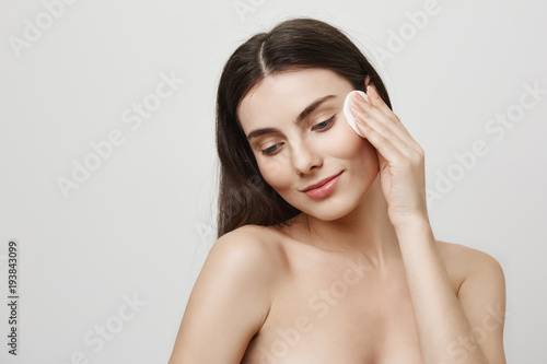 Pursuing beauty. Indoor shot of charming young european female standing in bathroom while wiping skin with cotton pad and skincare treatment, posing in studio over gray background