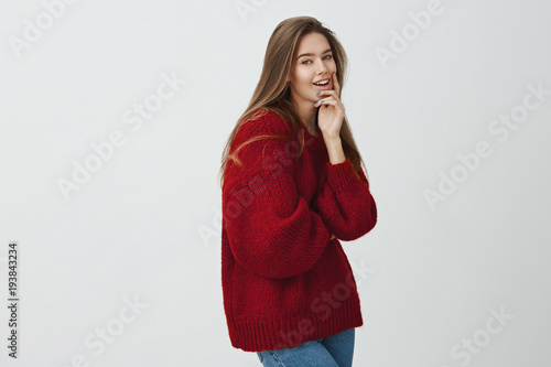 She is real heartbreaker. Indoor shot of attractive female student standing in profile, smiling at camera while touching chin and thinking about something, being curious if plan will be fulfilled © Cookie Studio