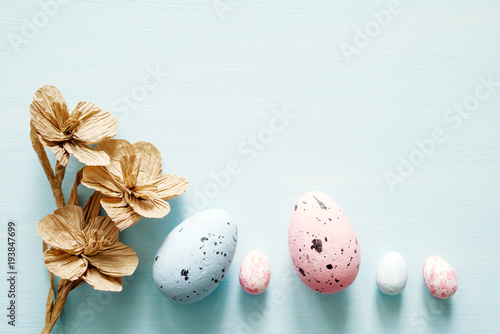 Easter eggs and decorative paper flowers on light blue background, top view, copy space