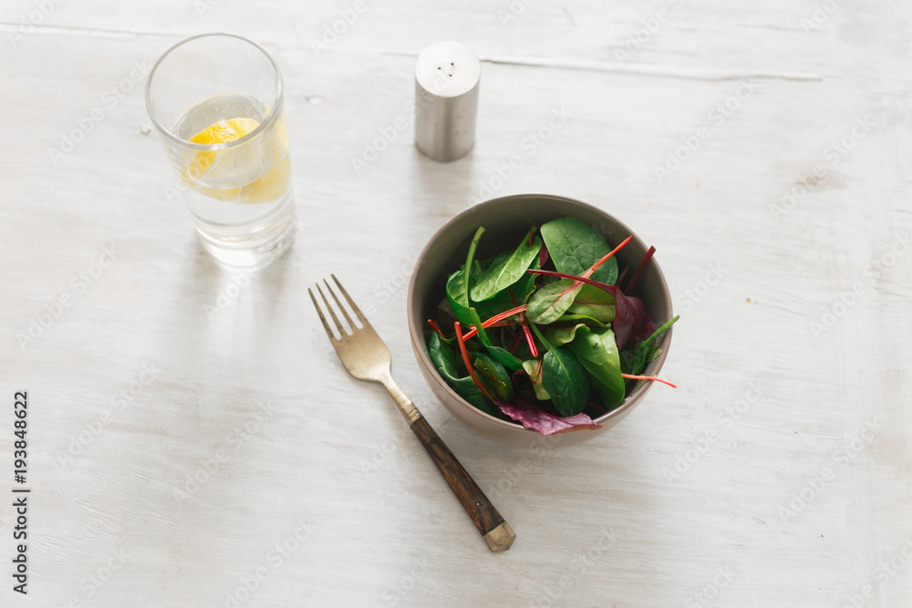 Vegetarian biodynamic food concept. Bowl of salad with spinach leaves and beet leaves on white wooden table with glass of water with lemon. Top view Vegetarian biodynamic food concept. Bowl of salad with spinach leaves and beet leaves on white wooden table with glass of water with lemon. Top view