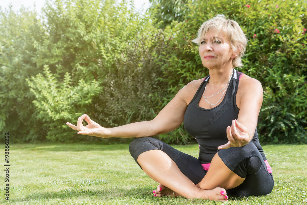Attractive senior blond woman is sitting cross legged doing yoga in the garden. 