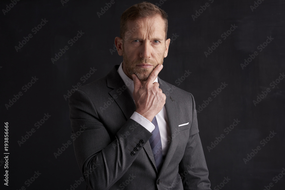 Thinking businessman portrait. Studio shot of wrinkled face business man wearing suit and looking thoughtful while sitting at dark background. Professional man wearing suit and tie. Thinking businessman portrait. Studio shot of wrinkled face business man wearing suit and looking thoughtful while sitting at dark background. Professional man wearing suit and tie.