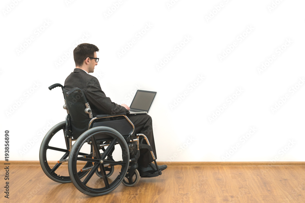 The happy disabled in a wheelchair works with a laptop on a white wall background The happy disabled in a wheelchair works with a laptop on a white wall background