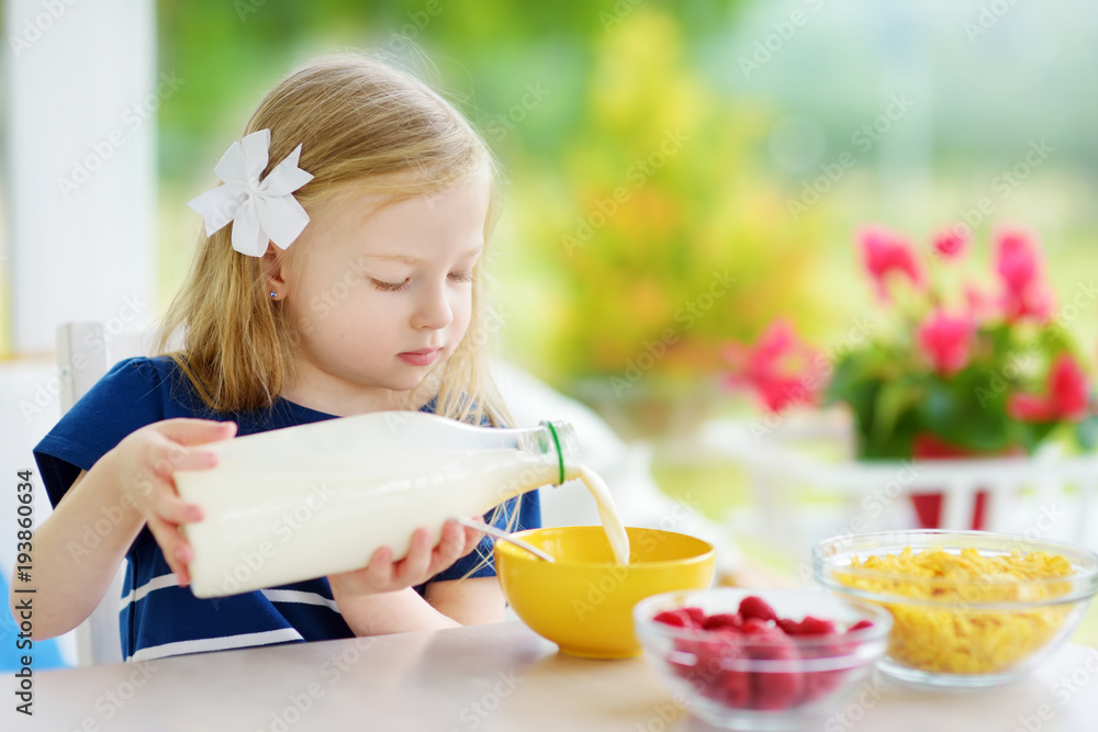 Cute little girl enjoying her breakfast at home. Pretty child eating corn flakes and raspberries and drinking milk before school.