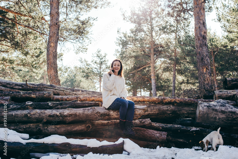 Fototapeta premium Young and pretty woman using her mobile phone outdoors, having fun taking herself pictures on a winter day with seated in a wooden trunk. Lifestyle.