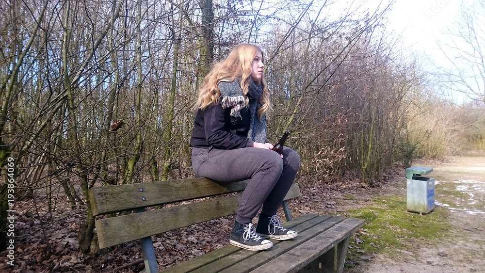 Fototapeta premium Teenager sitzt mit einer Bierflasche auf der Lehne einer Parkbank, Seitenansicht
