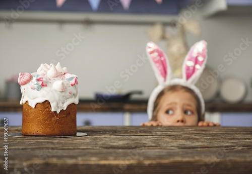 Canvas Print Cute little sweet tooth in bunny ears headband peeking out from behind kitchen table and looking at tasty easter cake with interest
