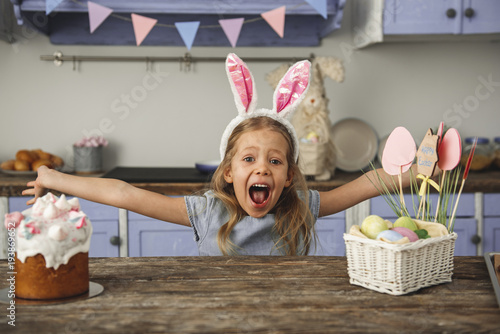Portrait of cheery little girl in bunny ears headband standing at kitchen table with easter cake and basket with eggs and screaming with outstretched arms