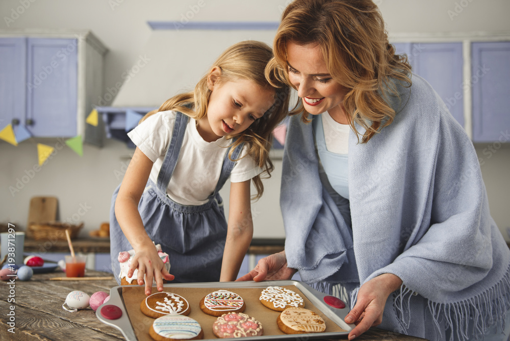 Fototapeta premium Satisfied mother and daughter standing in the kitchen and looking at cookies prepared for easter