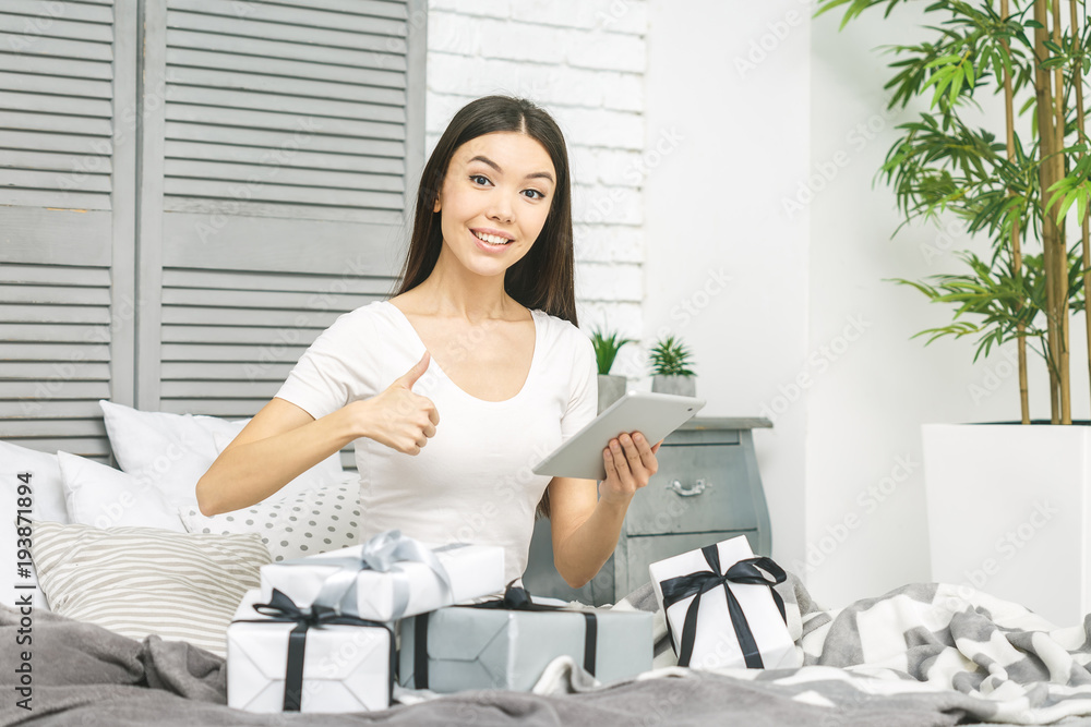 Smiling happy beautiful woman with gift boxes lying in bed and using tablet computer. Online shopping. Packing gifts. Thumbs up woman Smiling happy beautiful woman with gift boxes lying in bed and using tablet computer. Online shopping. Packing gifts. Thumbs up woman