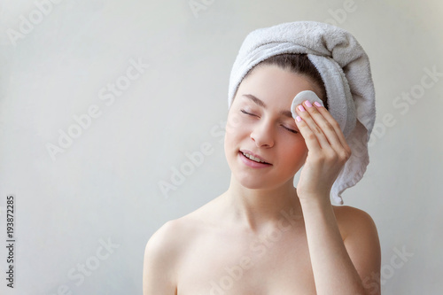 Beauty portrait of a smiling brunette woman in towel on head with soft healthy skin removing make up with cotton pad isolated over white background. Skincare cleansing spa relax concept